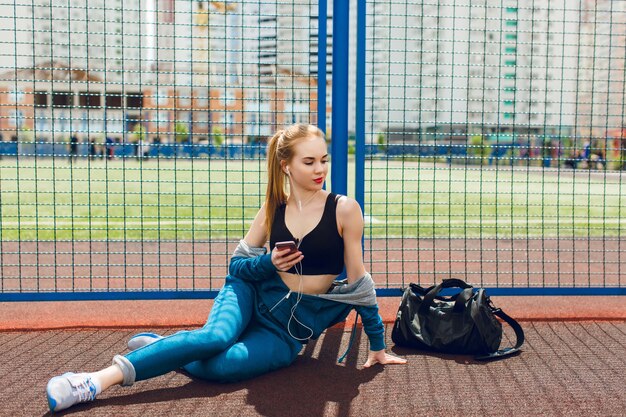 Una joven con un traje deportivo azul con un top negro está sentada cerca de la valla del estadio. Ella escucha la música con auriculares y mira a lo lejos.