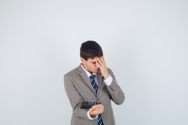 Joven en traje formal sosteniendo la calculadora, sosteniendo la mano en la frente y mirando molesto