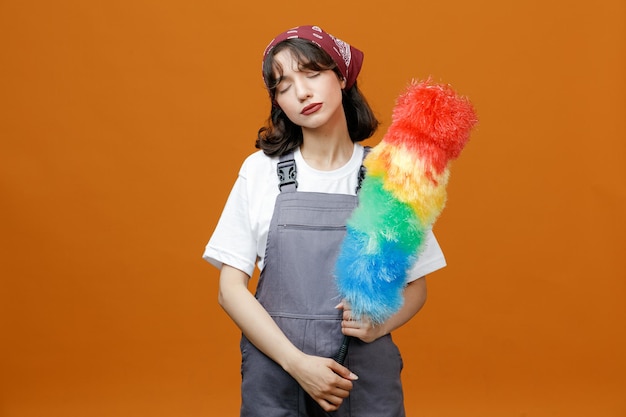 Foto gratuita una joven y tranquila limpiadora con uniforme y pañuelo sosteniendo un plumero con ambas manos con los ojos cerrados aislados en un fondo naranja