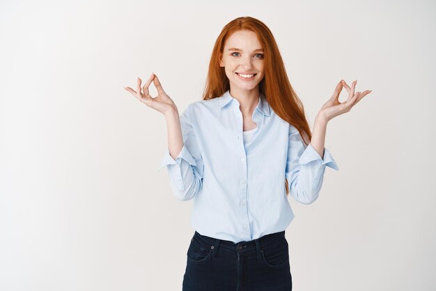 Joven tranquila mujer de negocios con cabello rojo meditando, tomados de la mano en el signo de mudra zen y sonriendo pacíficamente, de pie relajado contra la pared blanca