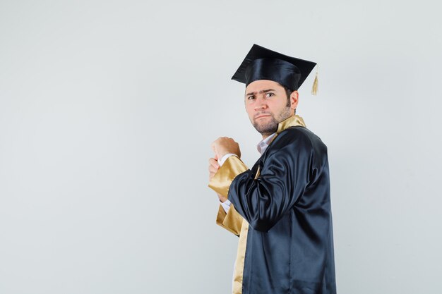 Joven tratando de abrir una botella de píldoras en uniforme de posgrado y con curiosidad.