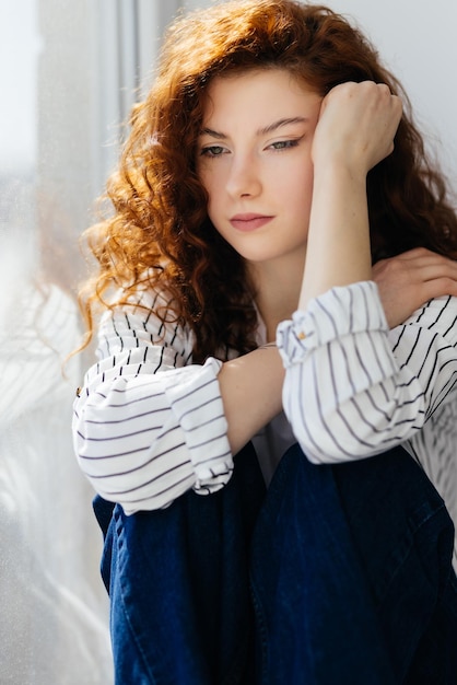 Foto gratuita una joven triste con el pelo rojo está sentada en el alféizar de la ventana