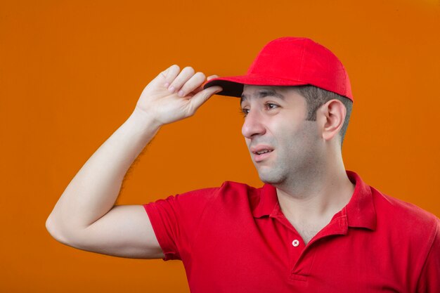 Joven triste repartidor molesto en polo rojo y gorra mirando a un lado i sobre fondo naranja aislado