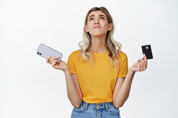 Una joven triste sosteniendo un teléfono móvil y una tarjeta de crédito mirando hacia arriba molesta por ser pobre y sin dinero en una cuenta bancaria de pie sobre fondo blanco