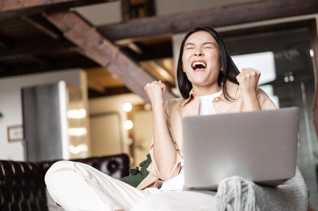 Joven triunfante asiática ganando reaccionando feliz en la notificación de buenas noticias de la computadora portátil gritando sí