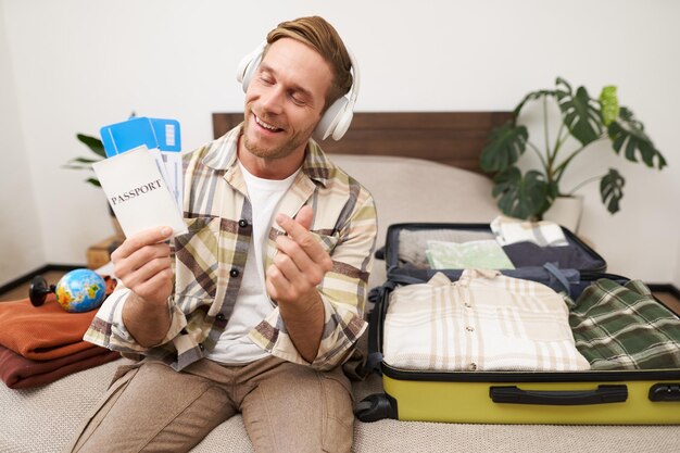 Un joven turista feliz muestra el pasaporte y los boletos va de vacaciones se sienta con la maleta empacada en la cama