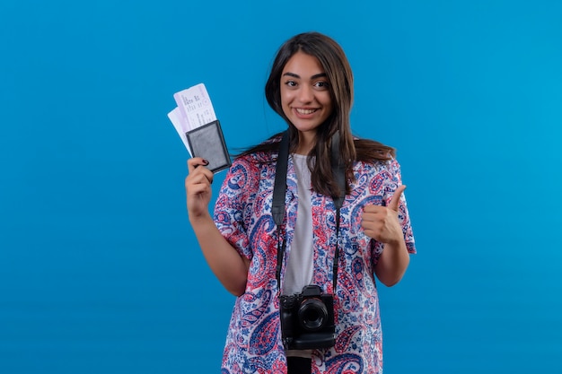 Joven turista hermosa mujer con cámara con pasaporte con boletos sonriendo alegremente, mostrando los pulgares para arriba, listo para vacaciones sobre pared azul aislado