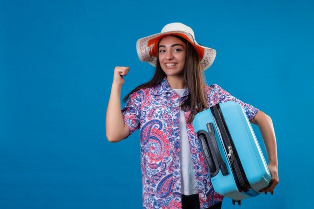 Joven turista hermosa mujer con sombrero de verano con maleta de viaje positiva y feliz levantando el puño después de una victoria, sonriendo alegremente, listo para viajar sobre bl aislado