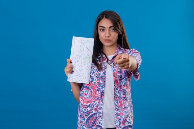 Joven turista hermosa mujer sosteniendo un mapa apuntando a la cámara con el dedo índice con expresión seria segura en la cara sobre la pared azul aislada