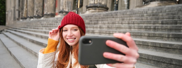 Joven turista pelirroja se toma un selfie frente al museo en las escaleras sostiene un teléfono inteligente y mira el móvil