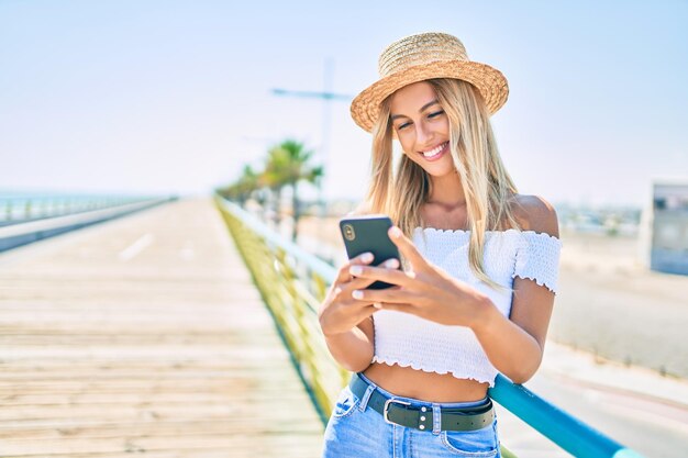 Joven turista rubia sonriendo feliz usando un smartphone en el paseo marítimo.