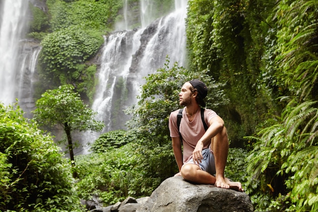 Joven turista varón caucásico descalzo con mochila sentado sobre una roca rodeada por la selva y admirando la hermosa vista con cascada