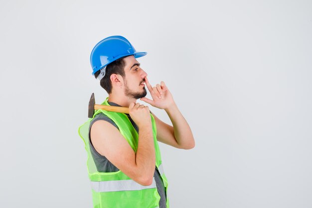 Joven en uniforme de construcción levantando el martillo sobre el hombro y mostrando gesto de silencio y mirando enfocado