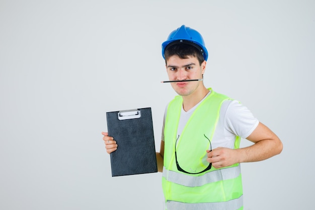 Foto gratuita joven en uniforme de construcción sosteniendo portapapeles y gafas de seguridad en las manos, sosteniendo la pluma con la boca y mirando serio