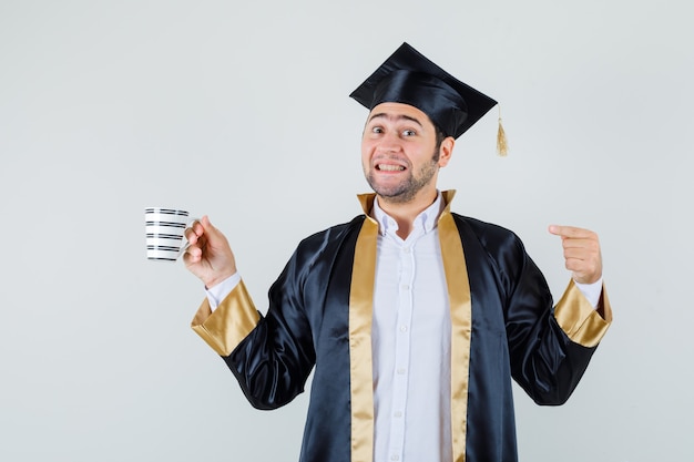 Joven en uniforme graduado apuntando a la taza de café y mirando alegre, vista frontal.