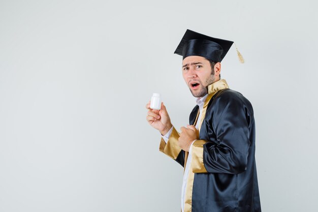 Joven en uniforme graduado sosteniendo una botella abierta de píldoras y mirando disgustado.