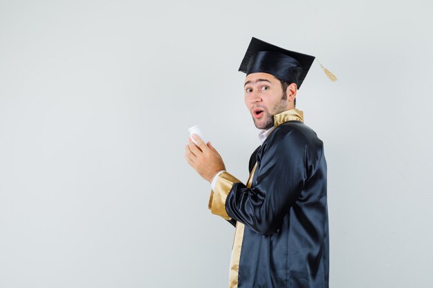 Joven en uniforme graduado sosteniendo una botella de píldoras y mirando asombrado