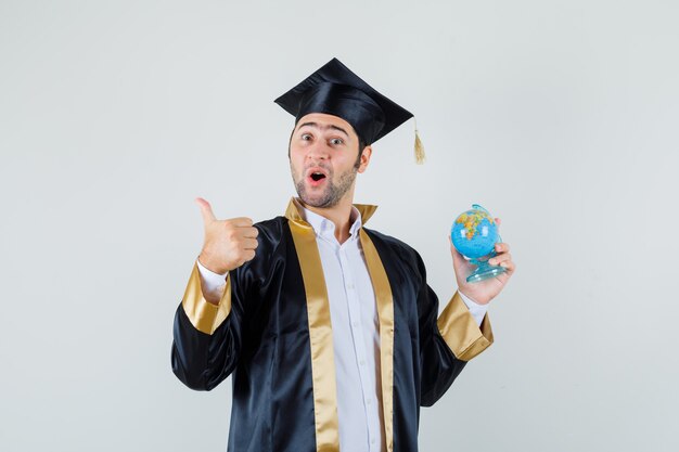 Joven en uniforme graduado sosteniendo el globo de la escuela, mostrando el pulgar hacia arriba y mirando alegre, vista frontal.