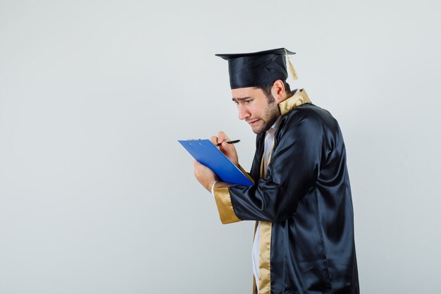 Joven en uniforme graduado tomando notas en el portapapeles y mirando triste.