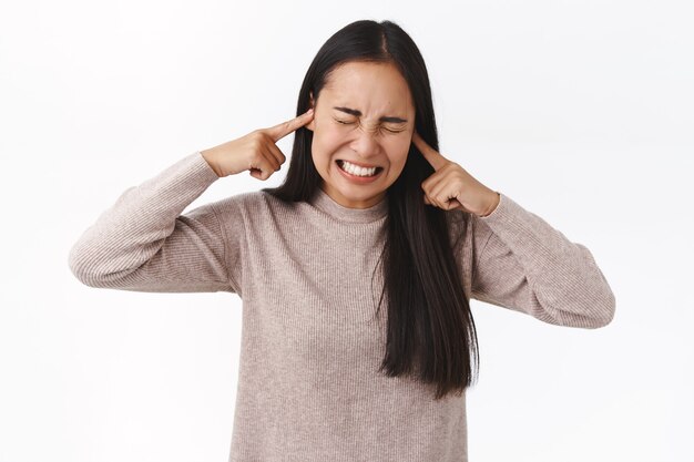 La joven universitaria asiática perturbada y molesta no puede estudiar, preparar exámenes, causar ruido, vecinos vomitar, cerrar las orejas con los dedos, hacer muecas de angustia