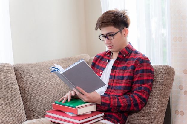 Joven usando anteojos sentado en el sofá, sonríe con alegría y mirando un libro en la mano