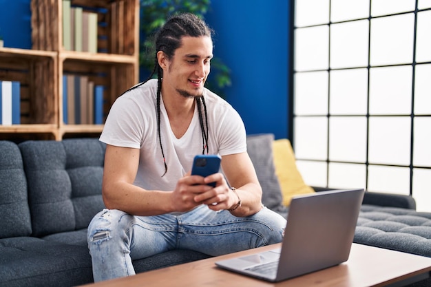 Joven usando un teléfono inteligente y una computadora portátil sentado en un sofá en casa