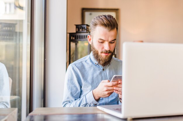 Foto gratuita joven, utilizar, teléfono celular, con, computador portatil, en el escritorio