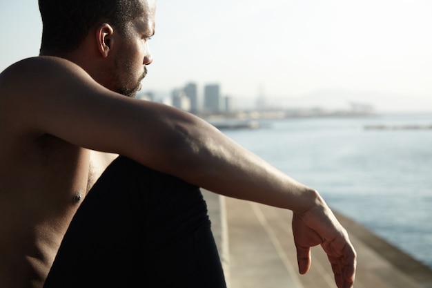 Joven vagabundo sentado en el paseo del río con el brazo estirado descansando sobre su rodilla. El hombre de piel negra está pensando en su vida en la gran ciudad y observa las olas de agua, relajándose bajo la luz del sol.