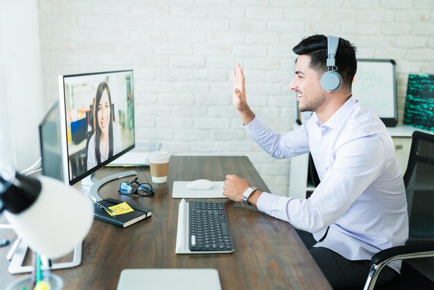 Joven vendedor confiado saludando a su colega durante una videollamada en la computadora en el escritorio