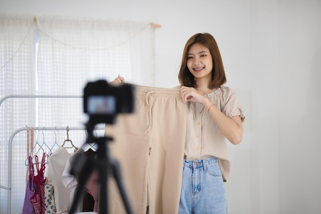 Foto gratuita joven vendedora asiática que muestra ropa de mujer y hace video en vivo en línea en casa mercado en línea con concepto de redes sociales