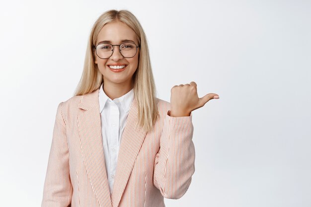 Joven vendedora de gafas y traje, apuntando con el dedo hacia la derecha, sonriendo en blanco