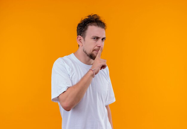 Joven vestido con camiseta blanca mostrando gesto de silencio en la pared naranja aislada