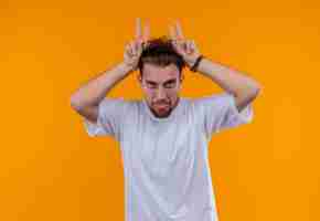 Foto gratuita joven vestido con camiseta blanca mostrando la lengua y el gesto de orejas de conejo en la pared naranja aislada