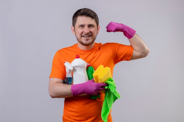 Joven vestido con camiseta naranja y guantes de goma, herramientas de limpieza sonriendo alegremente positivo y feliz mostrando bíceps