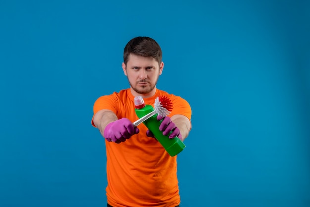 Joven vestido con camiseta naranja y guantes de goma con productos de limpieza y cepillo para fregar