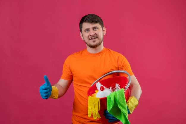 Joven vestido con camiseta naranja y guantes de goma sosteniendo un balde con herramientas de limpieza mirando confiado sonriendo positivo y feliz mostrando los pulgares para arriba de pie sobre la pared rosa