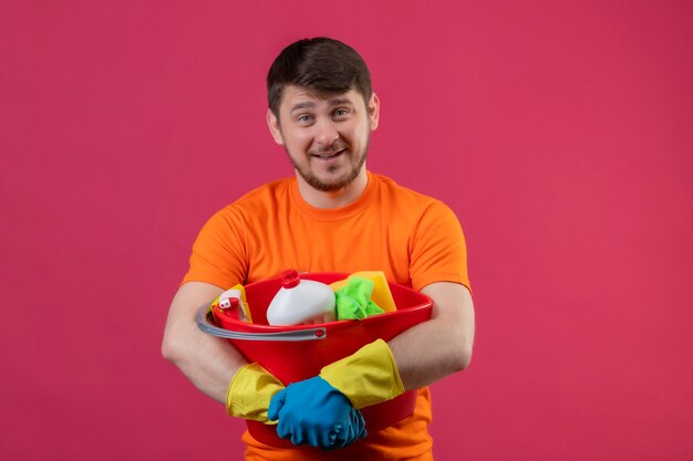 Joven vestido con camiseta naranja y guantes de goma sosteniendo un balde con herramientas de limpieza que parece seguro