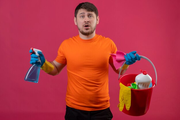 Joven vestido con camiseta naranja y guantes de goma sosteniendo un balde con herramientas de limpieza y spray de limpieza