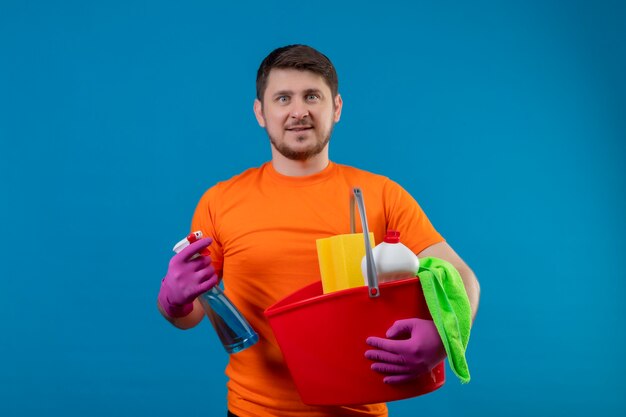 Joven vestido con camiseta naranja y guantes de goma sosteniendo un balde con herramientas de limpieza