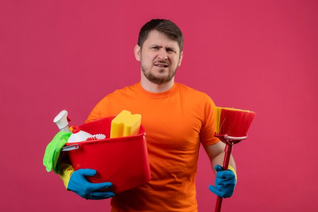 Joven vestido con camiseta naranja y guantes de goma sosteniendo un balde con herramientas de limpieza