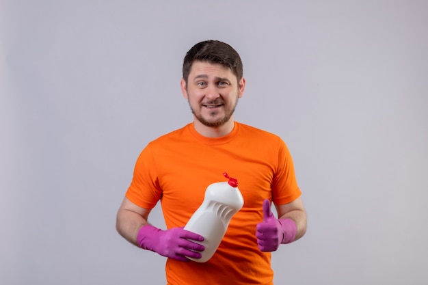 Foto gratuita joven vestido con camiseta naranja y guantes de goma sosteniendo una botella con productos de limpieza sonriendo de pie positivo y feliz sobre la pared blanca