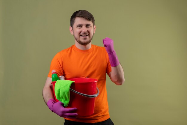 Joven vestido con camiseta naranja y guantes de goma sosteniendo un cubo con herramientas de limpieza apretando el puño feliz y salió sonriendo alegremente regocijándose de su éxito de pie sobre la pared verde