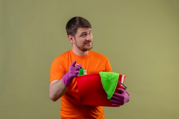 Joven vestido con camiseta naranja y guantes de goma sosteniendo un cubo con herramientas de limpieza sonriendo apuntando con el dedo a la cámara de pie feliz y positivo sobre la pared verde