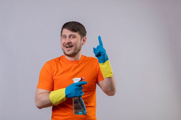 Joven vestido con camiseta naranja y guantes de goma sosteniendo spray de limpieza sonriendo alegremente