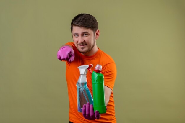 Joven vestido con camiseta naranja y guantes de goma sosteniendo suministros de limpieza sonriendo positivo y feliz apuntando con el dedo a la cámara de pie sobre la pared verde 2