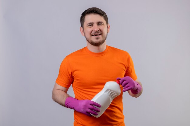 Joven vestido con camiseta naranja y guantes de goma sosteniendo suministros de limpieza sonriendo positivo y feliz de pie sobre la pared blanca