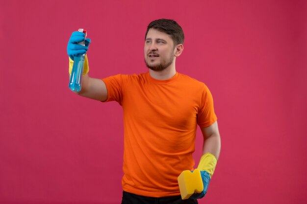 Joven vestido con camiseta naranja y guantes de goma con spray de limpieza y alfombra mirando confiado de pie sobre la pared rosa