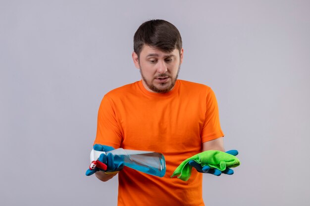 Joven vestido con camiseta naranja y guantes de goma con spray de limpieza y alfombra mirándolos