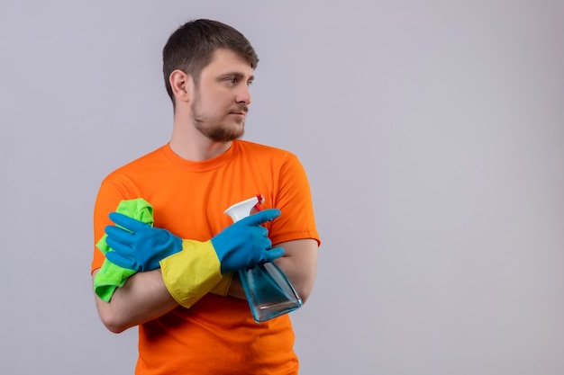 Joven vestido con camiseta naranja y guantes de goma con spray de limpieza y alfombra de pie con los brazos cruzados.