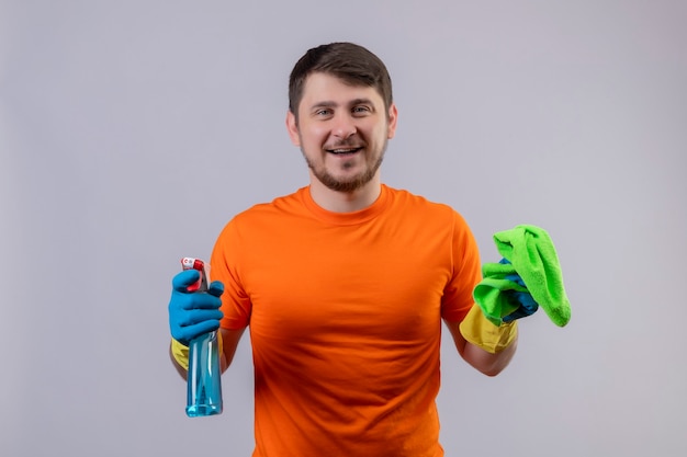 Foto gratuita joven vestido con camiseta naranja y guantes de goma con spray de limpieza y alfombra sonriendo alegremente positivo y feliz de pie sobre la pared blanca